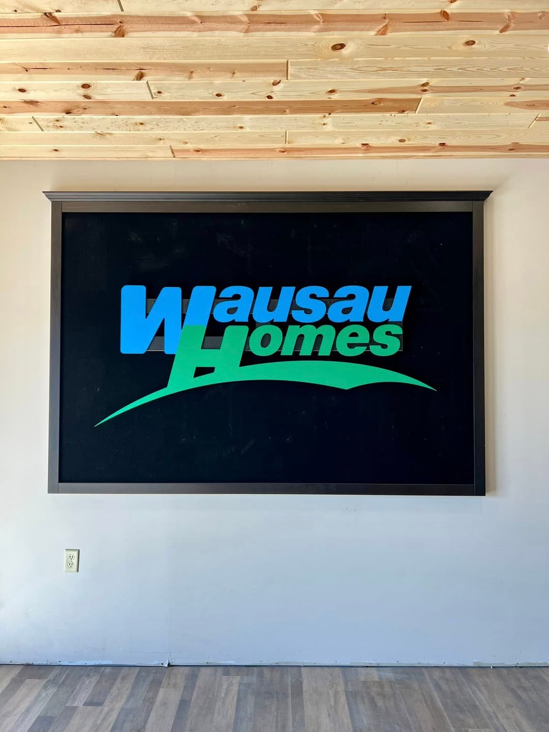 Wausau Homes logo displayed on a wall in an office setting with wooden ceiling.