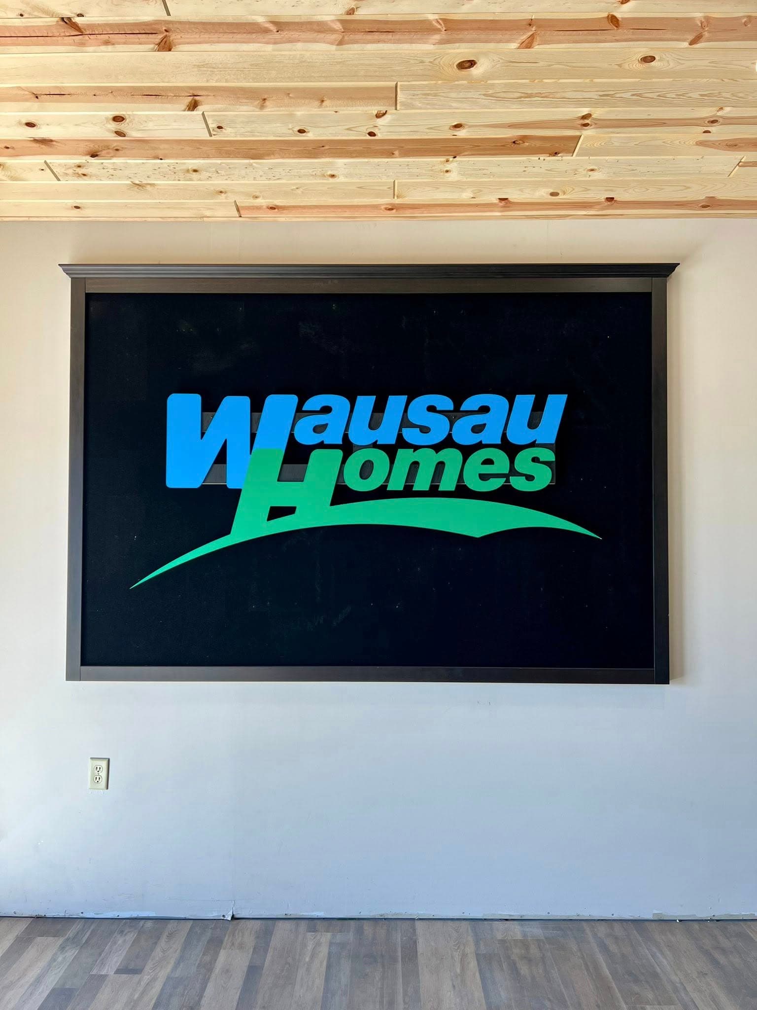 Wausau Homes logo displayed on a wall in an office setting with wooden ceiling.