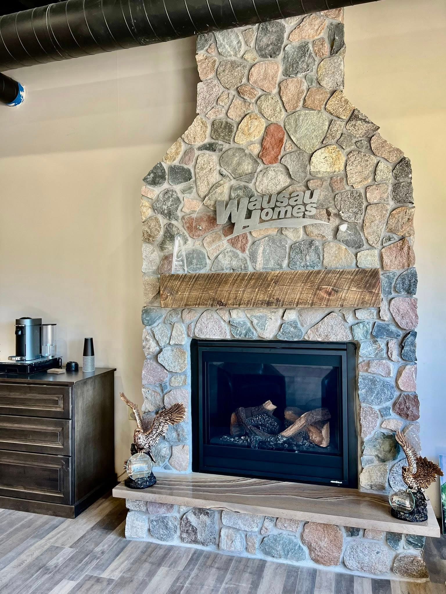 Stone fireplace with Wausau Homes logo, wooden mantel, and decorative accents in modern interior.