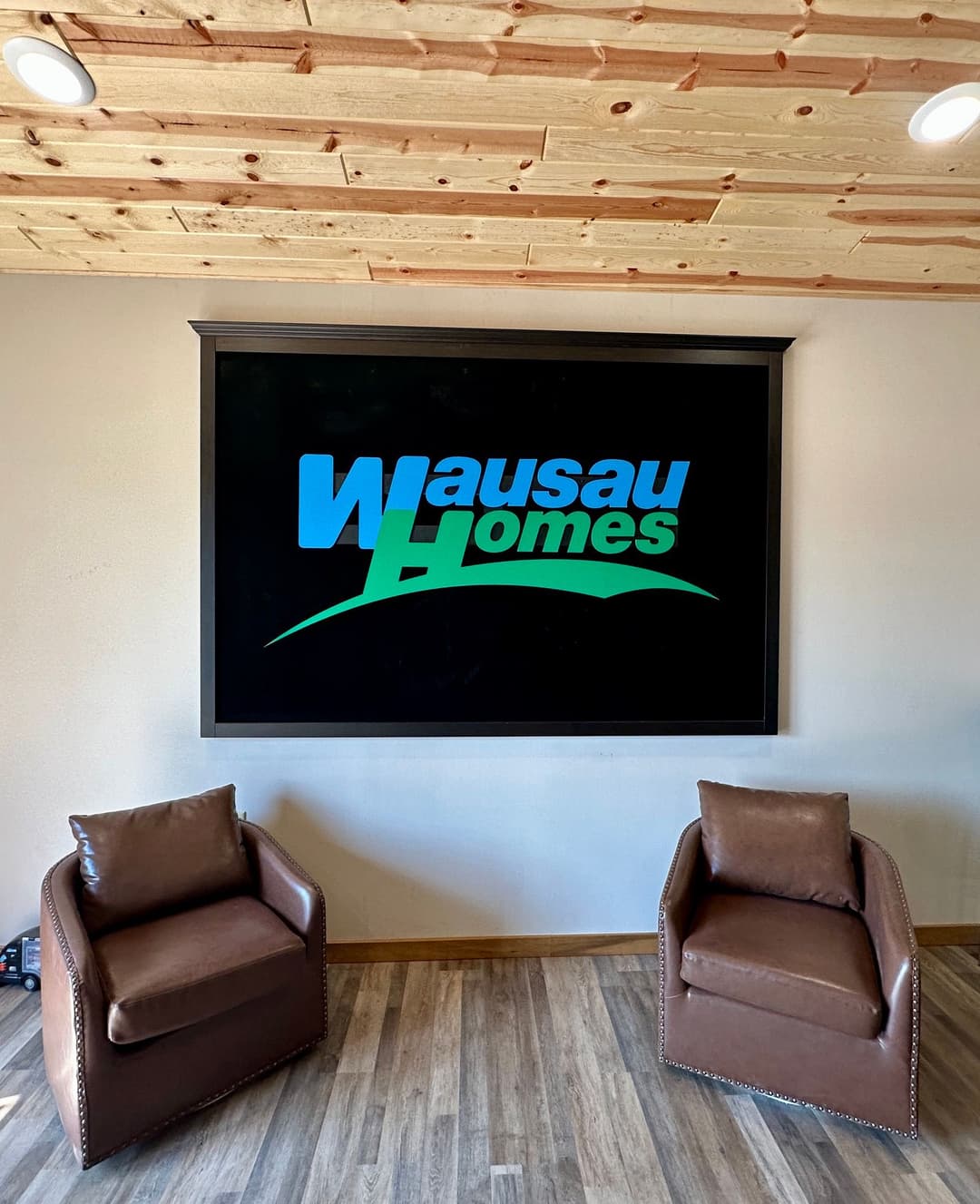 Wausau Homes logo displayed on a wall with two brown chairs in a modern interior setting.
